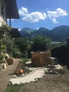Feuerwasser Badefass mit Außenofen im Garten vor Bergkulisse aus V4A-Edelstahl und Kiefernholz