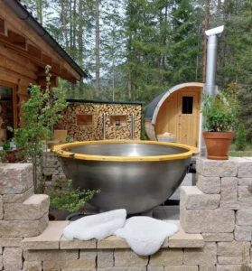 Badezuber aus Edelstahl und Kiefer-Vollholz von Feuerwasser auf einer Naturstein-Terrasse mit Sauna im Hintergrund