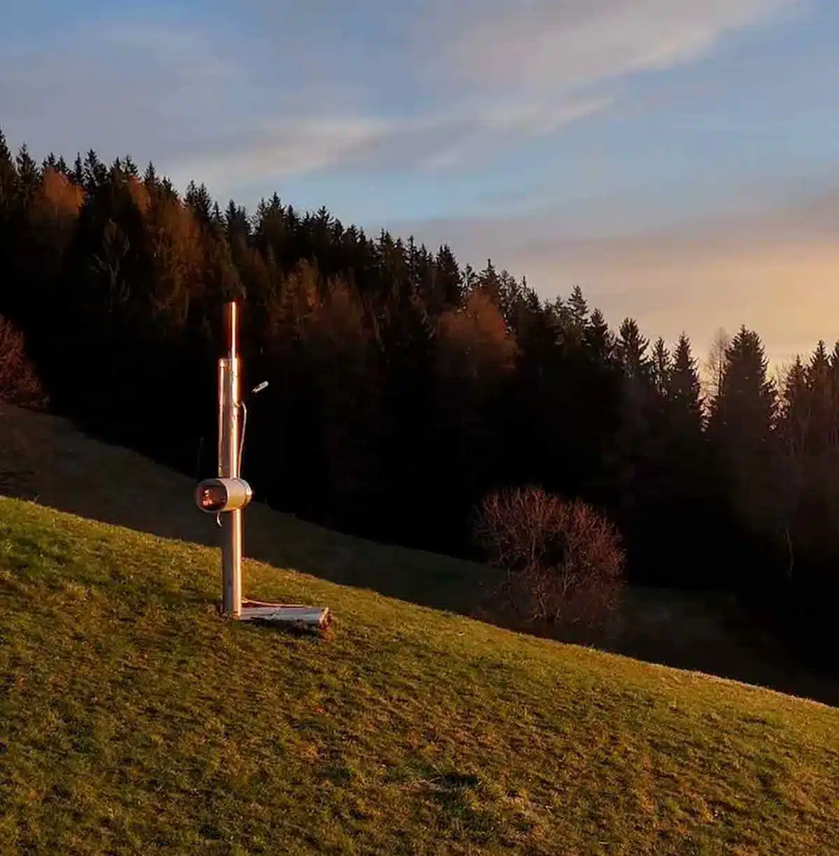 Holzbeheizte Gartendusche von Feuerwasser auf einer Hangwiese im warmen Licht des Sonnenuntergangs, umgeben von Wald