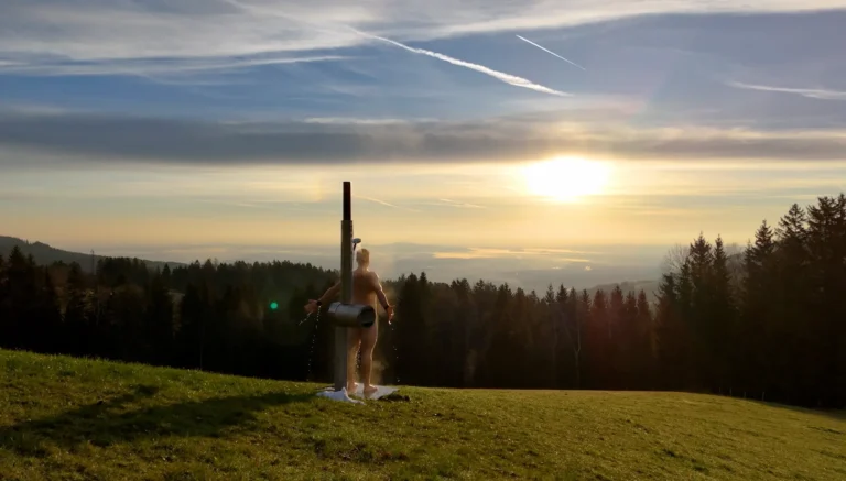 Firewater garden shower made of stainless steel on a mountain meadow at sunset, a man showers in the open air with a view over forests and valleys.