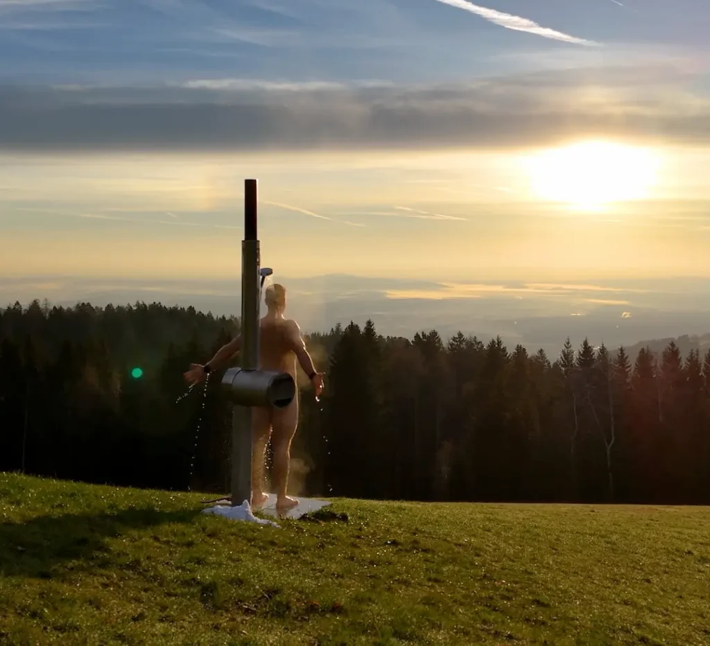 Feuerwasser Gartendusche aus Edelstahl auf einer Bergwiese bei Sonnenuntergang, ein Mann duscht in der freien Natur mit Blick über Wälder und Täler.