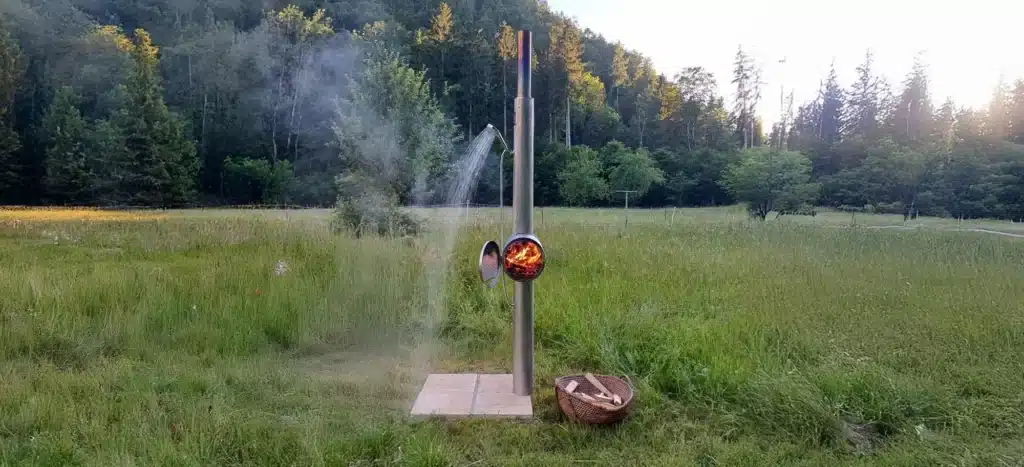 Gartendusche aus Edelstahl von Feuerwasser im Einsatz auf einer Wiese, mit brennendem Holzofen und fließendem warmem Wasser.