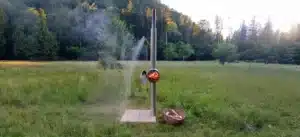Gartendusche aus Edelstahl von Feuerwasser im Einsatz auf einer Wiese, mit brennendem Holzofen und fließendem warmem Wasser.
