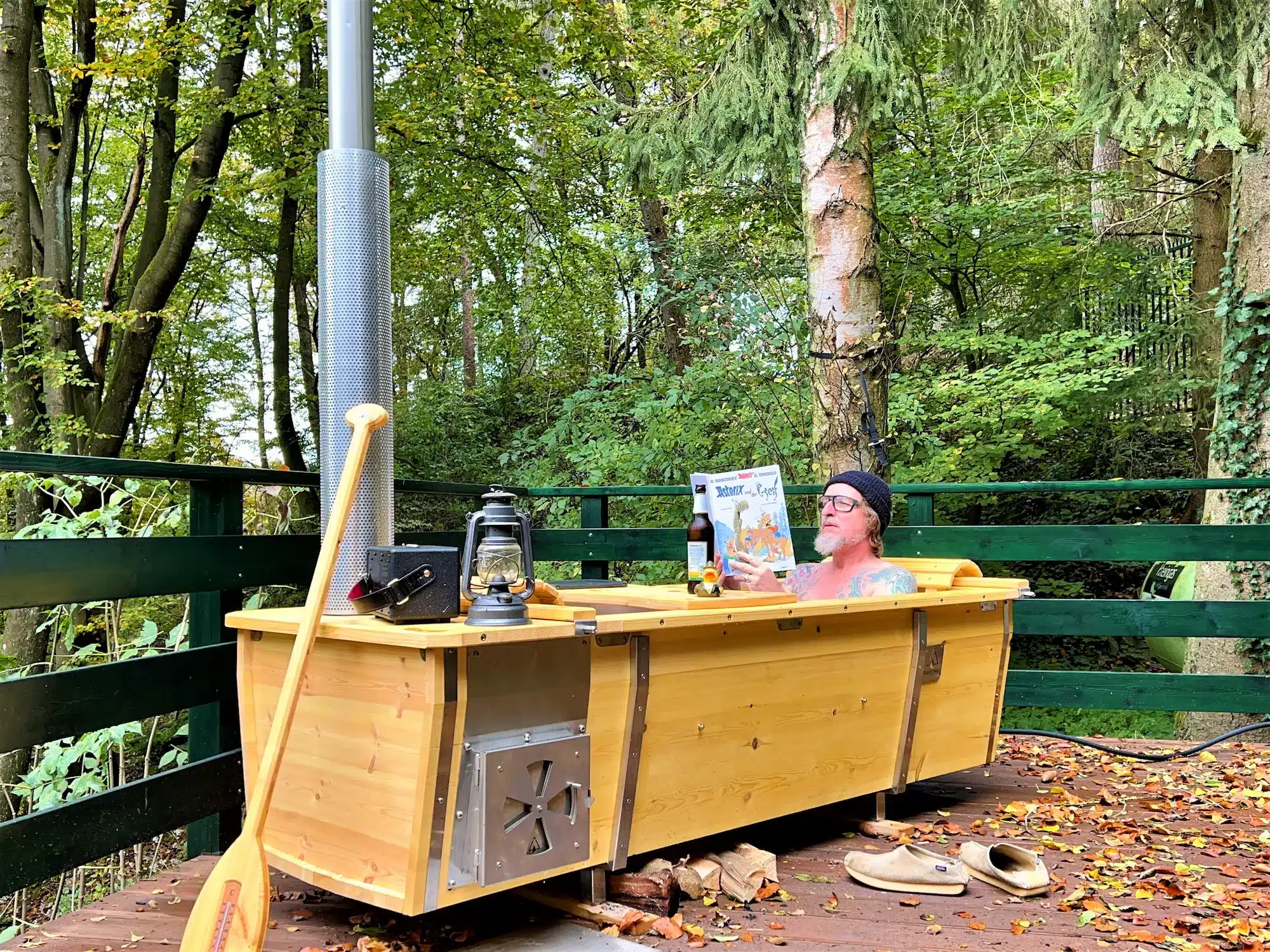 Outdoor-Badewanne aus V4A-Edelstahl mit Kiefernholz-Verkleidung auf Holzdeck im Wald, ein Mann badet und liest Asterix